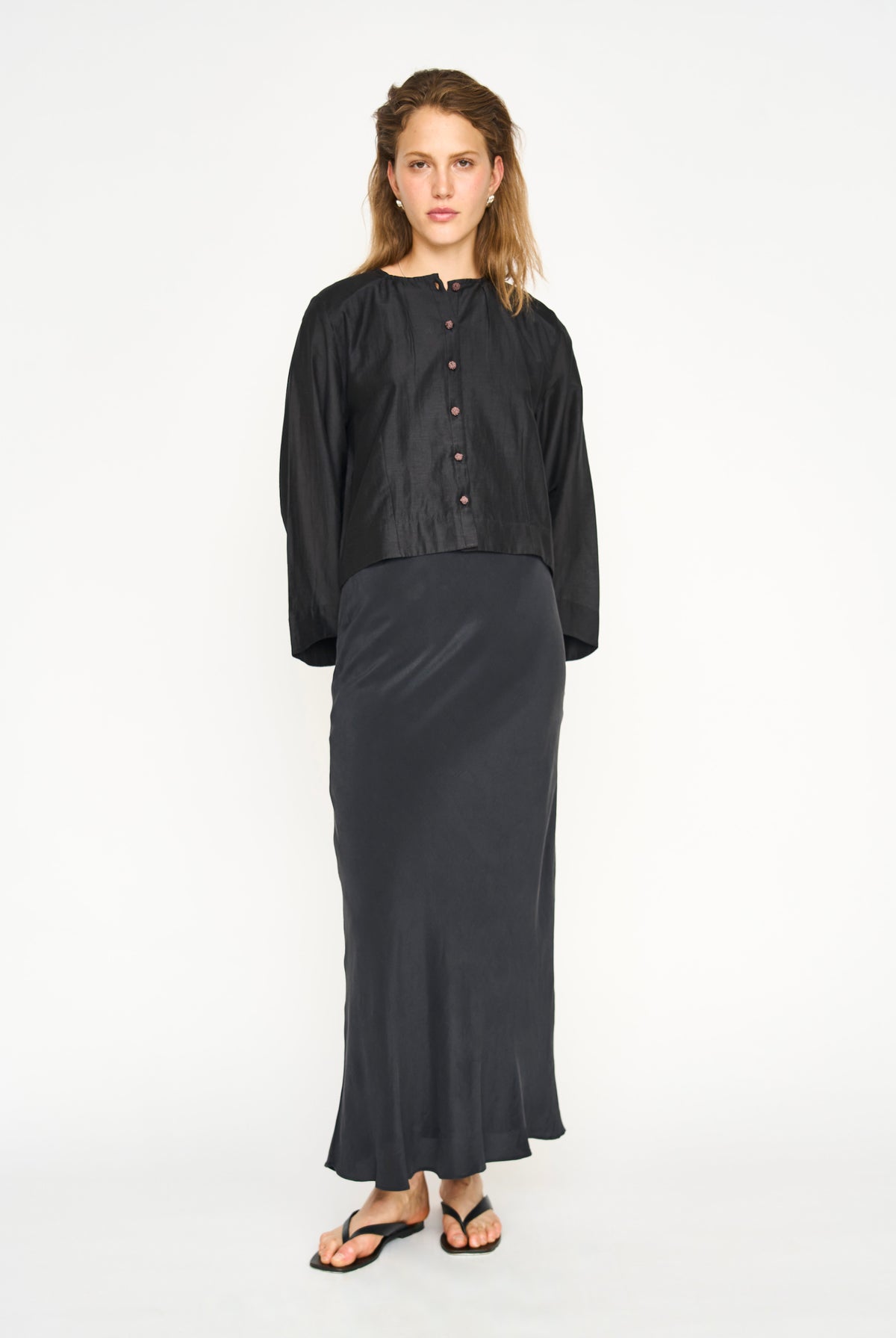 A woman modeling a black two-piece outfit consisting of a cropped long-sleeve button-front top and a matching long skirt, paired with black flip-flop sandals against a plain white studio background.