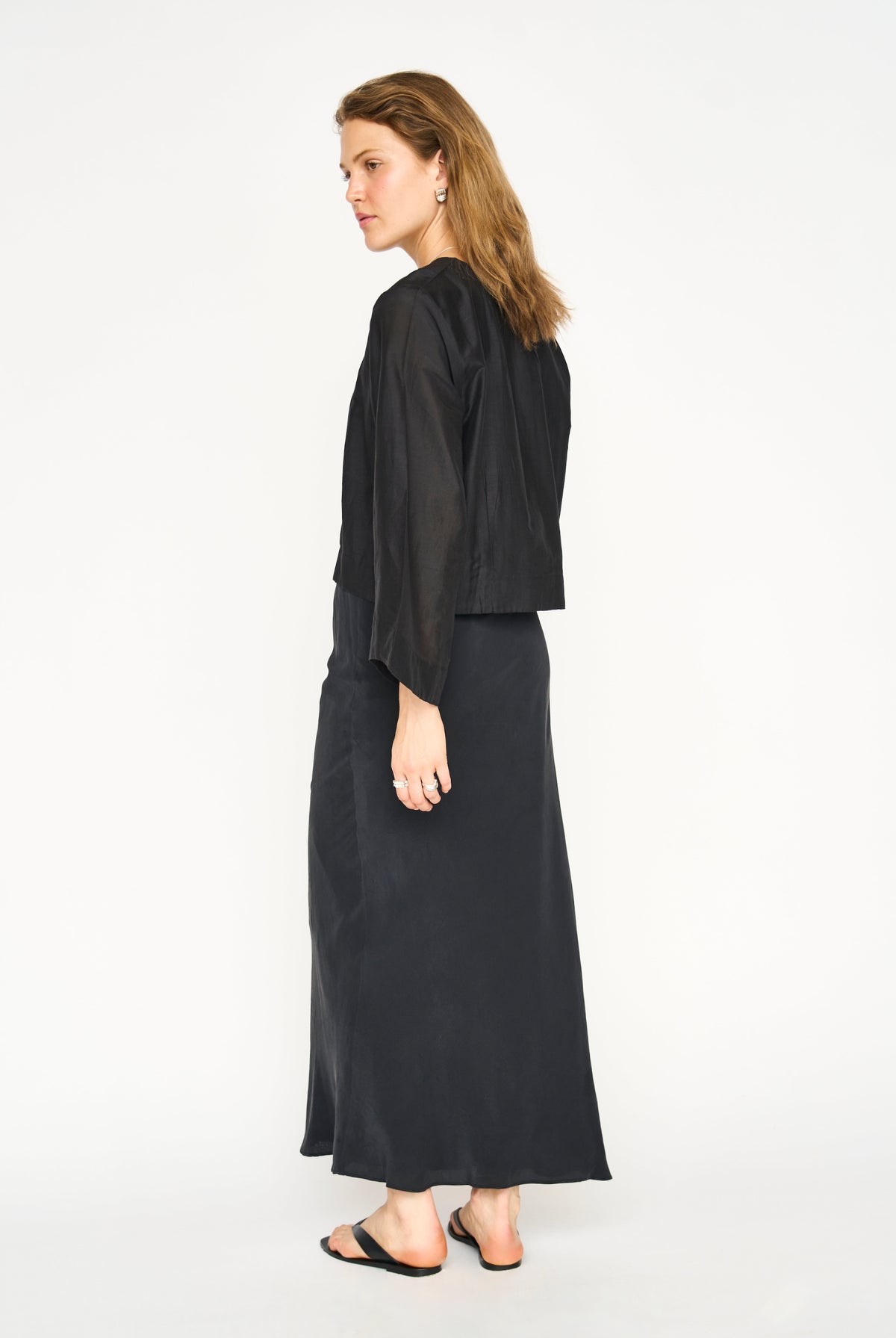 A woman modeling a black two-piece outfit consisting of a cropped long-sleeve top and a matching maxi skirt, shown from a side-back angle against a plain white studio background.