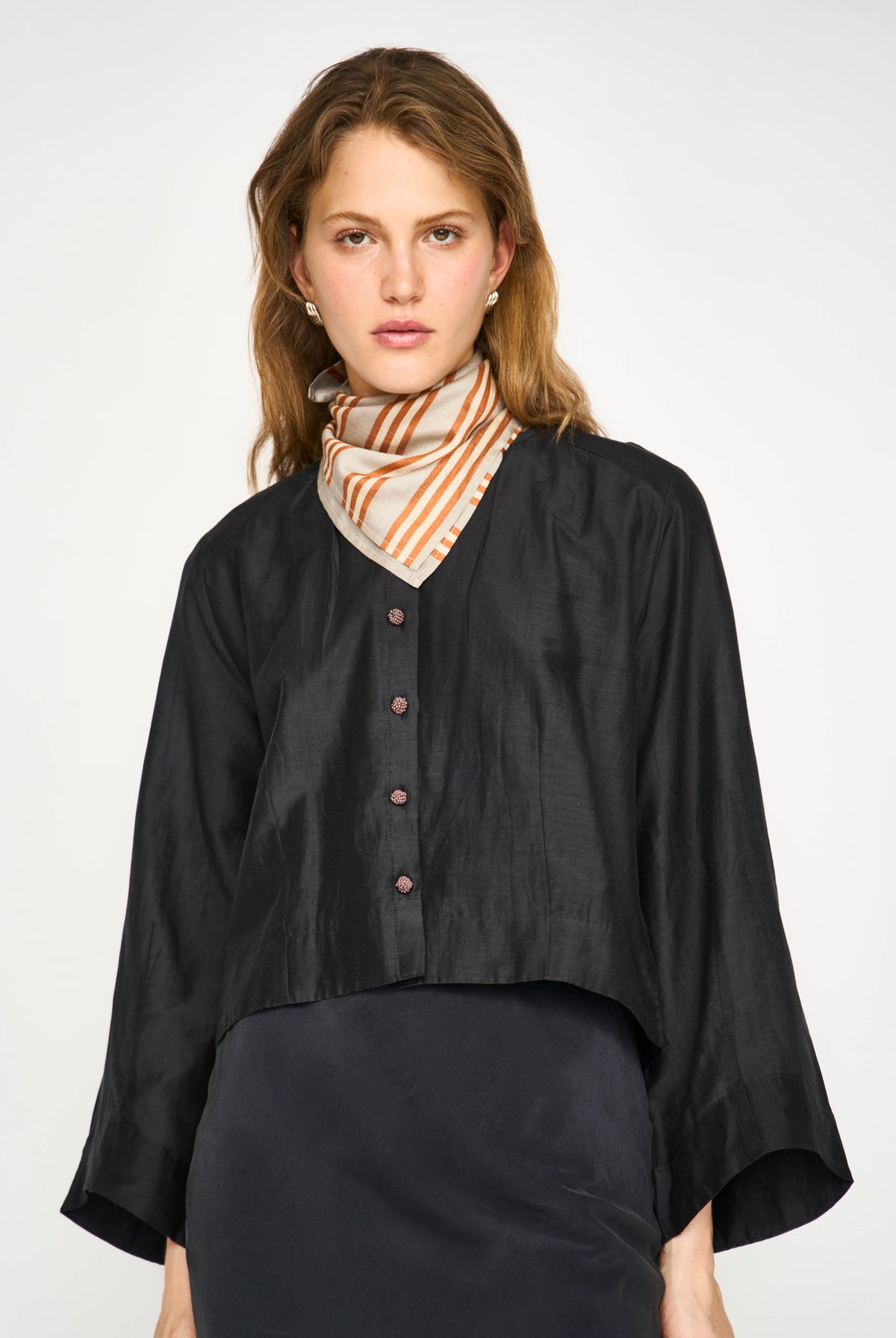 A woman modeling a black cropped button-front blouse with wide three-quarter sleeves, paired with a striped orange and white neck scarf, against a plain light gray studio background.
