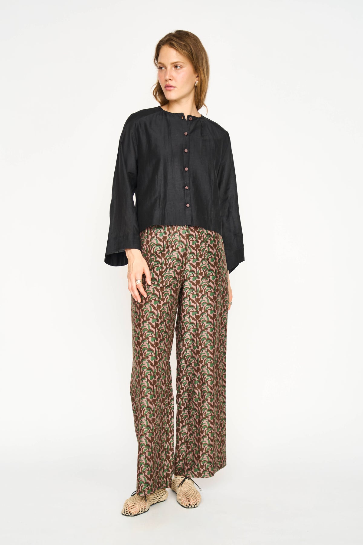 A woman modeling a black long-sleeve button-front blouse with wide cuffs, paired with brown and beige patterned wide-leg trousers, against a plain white studio background.