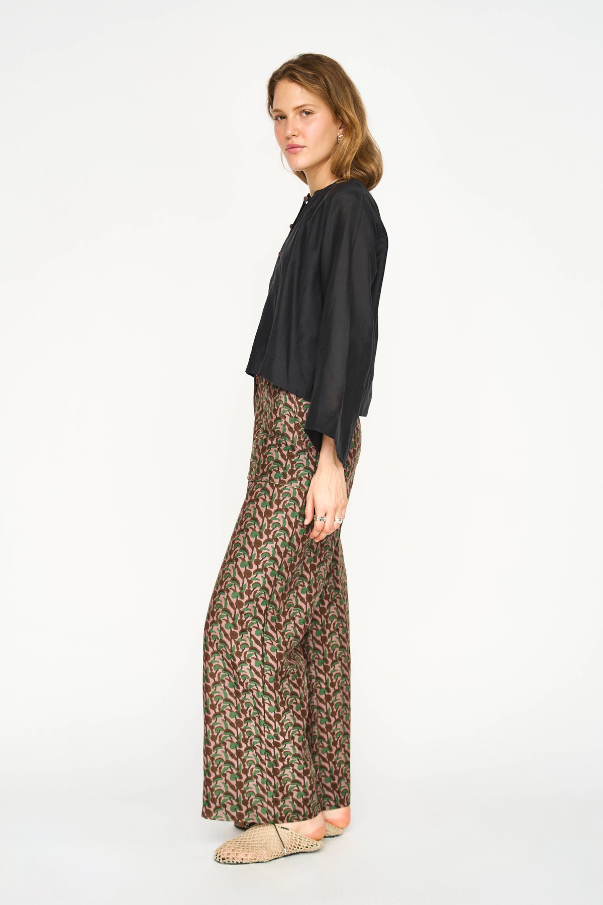 A woman modeling a black long-sleeve blouse and wide-leg patterned trousers with a beige woven slip-on shoe, photographed against a plain white studio background.