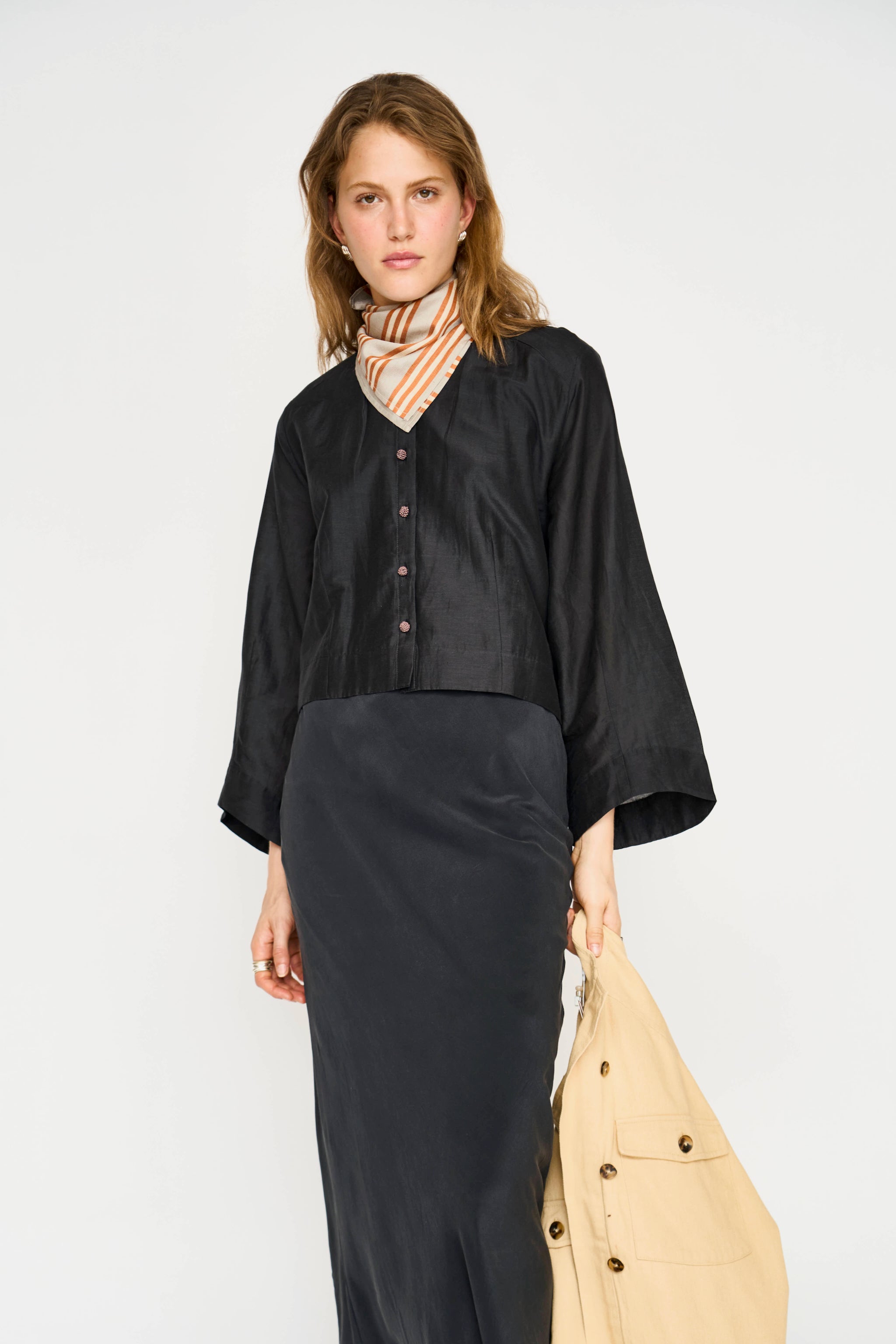A model stands against a light gray studio background wearing a black cropped button-front top with wide three-quarter sleeves, a striped neck scarf, a long black skirt, and holding a beige tote bag.
