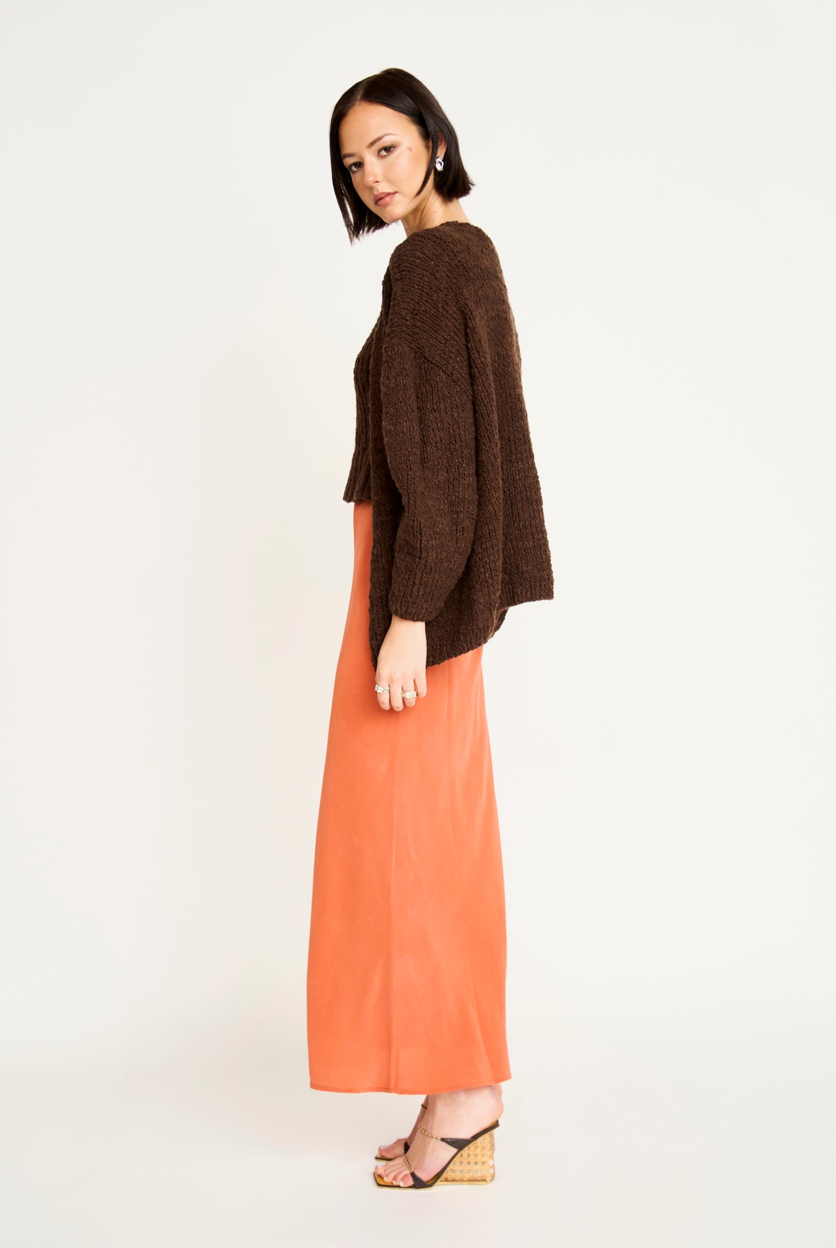 A model is shown from the side/back wearing a dark brown fuzzy knit cardigan and a long orange skirt, standing against a plain white studio background.