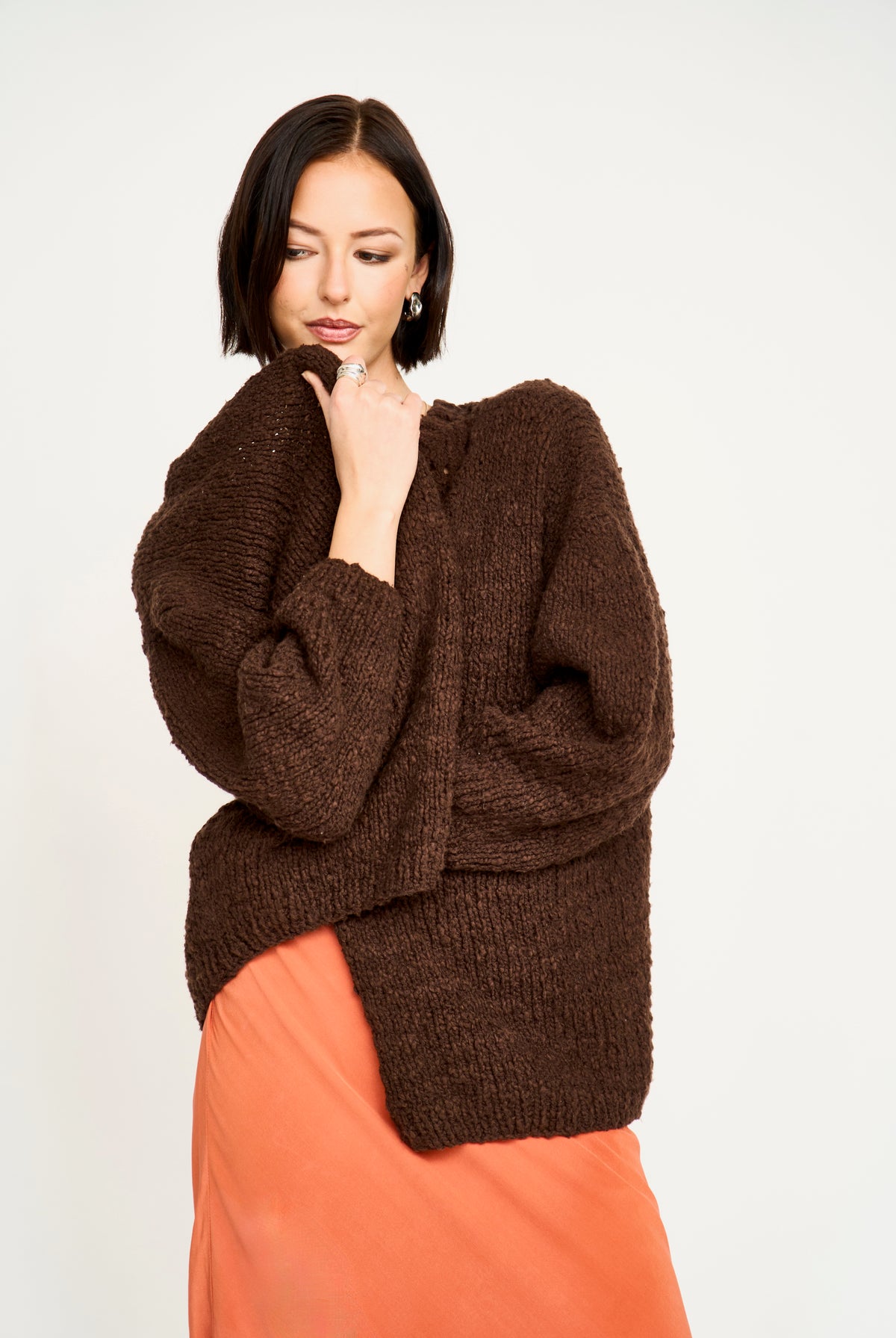 A woman wearing a chunky, oversized dark brown knit sweater and a peach-orange skirt, posing against a solid off-white studio background.
