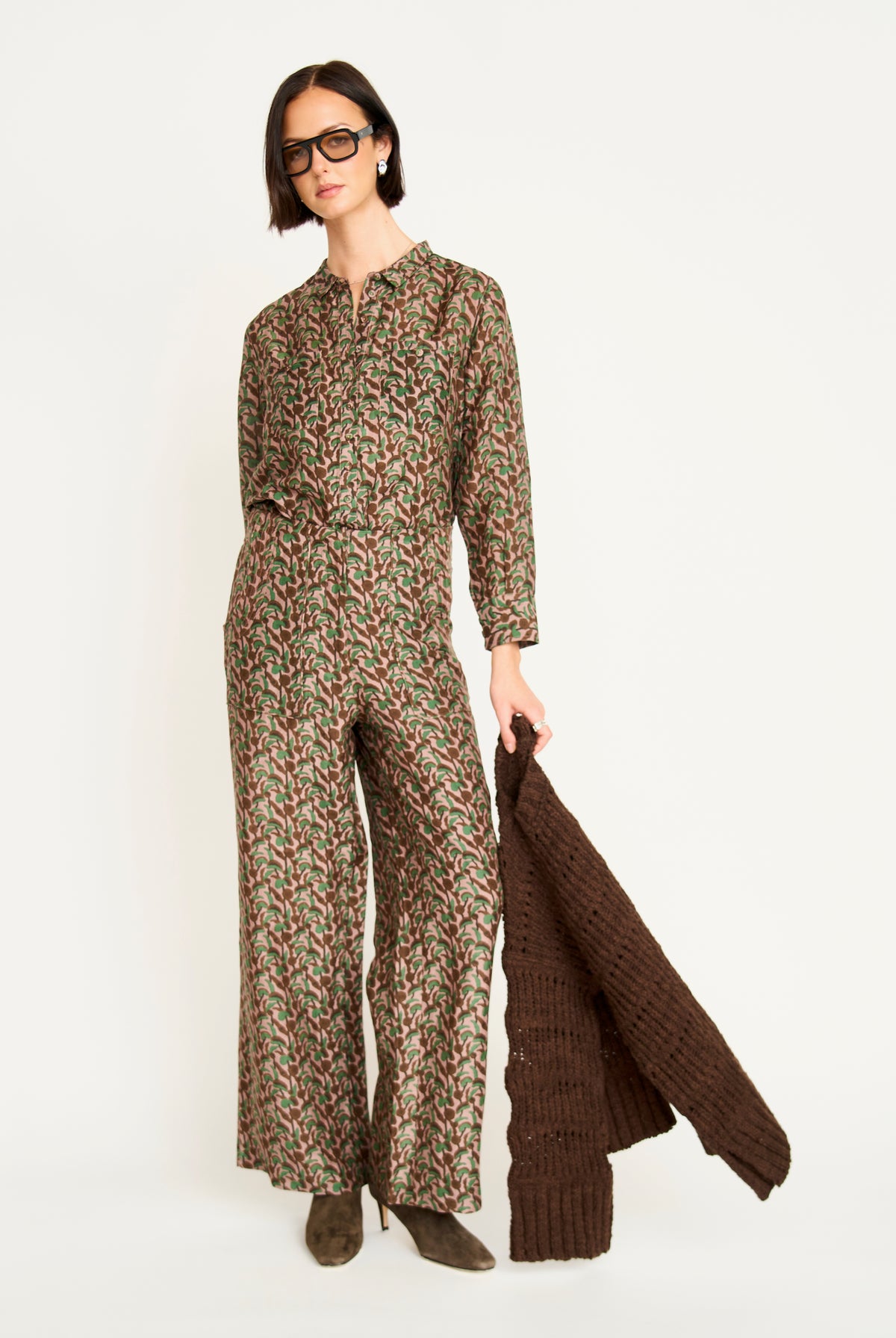 A model wearing a brown, green, and beige patterned wide-leg jumpsuit and holding a dark brown chunky knit cardigan against a plain white studio background.