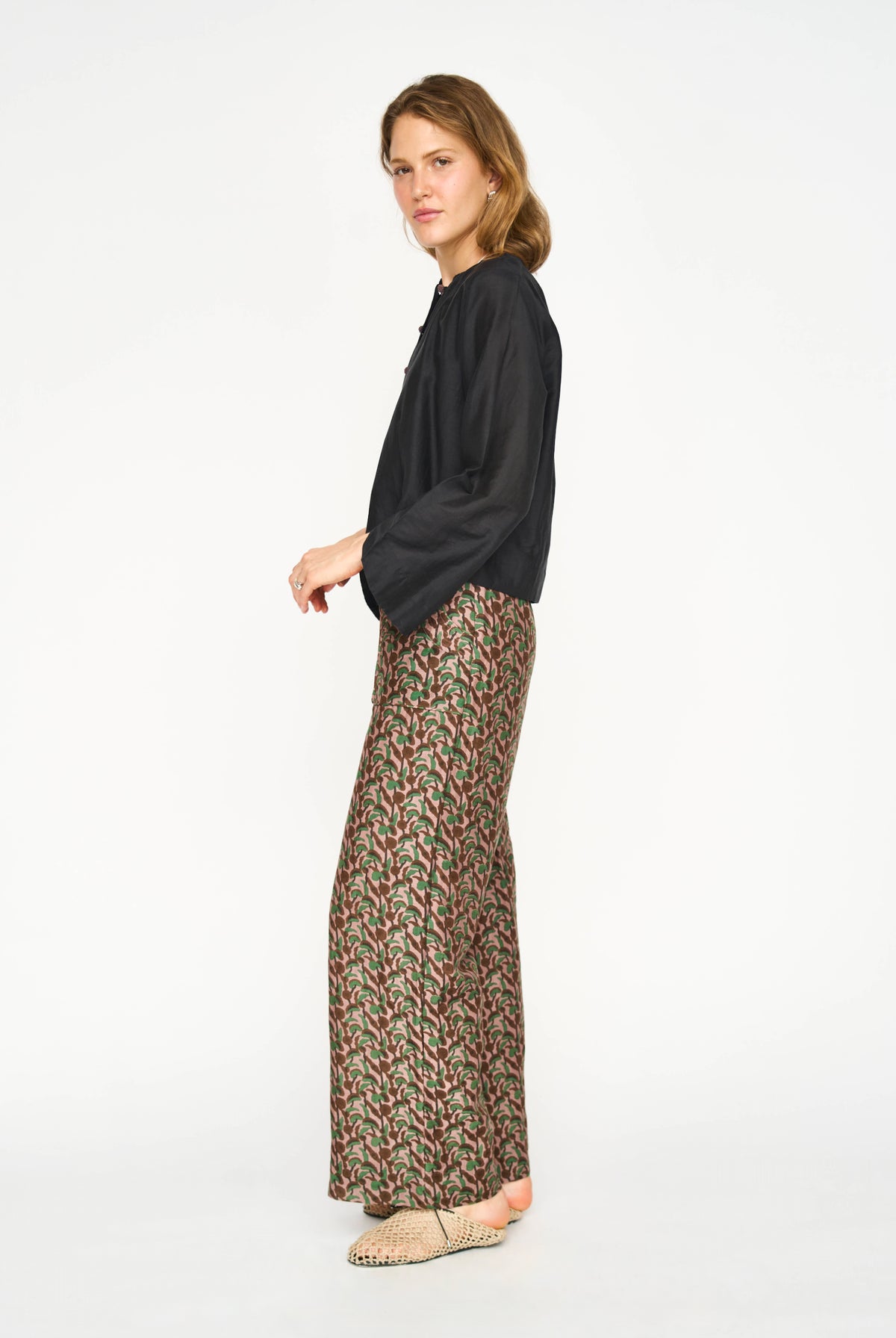 A woman modeling a black long-sleeve top and wide-leg trousers with a dense red, green, and beige geometric pattern, standing against a plain white studio background.