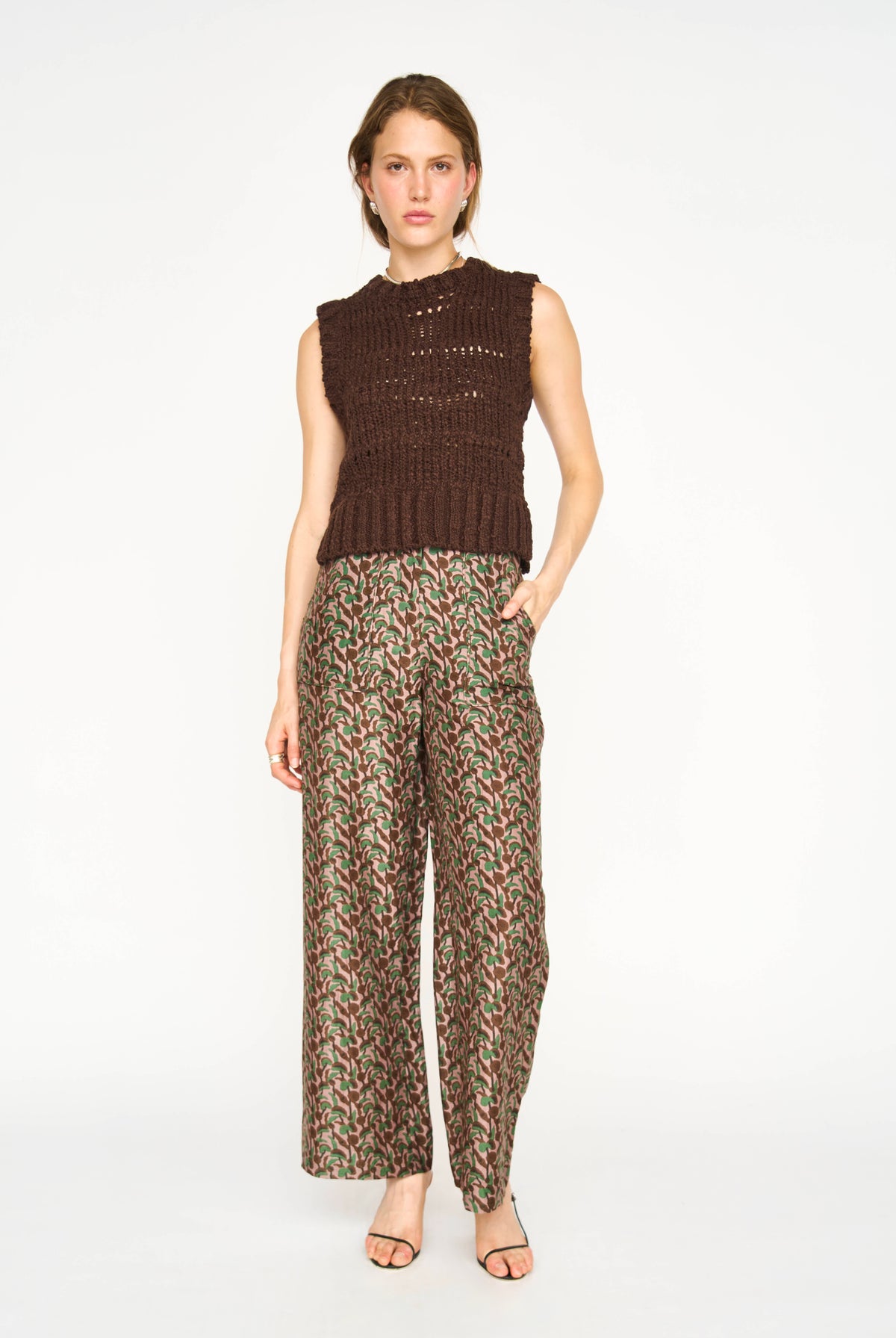 A model stands against a plain white studio background wearing a sleeveless dark brown knit top and wide-leg trousers with a dense red, green, and beige floral or abstract print.