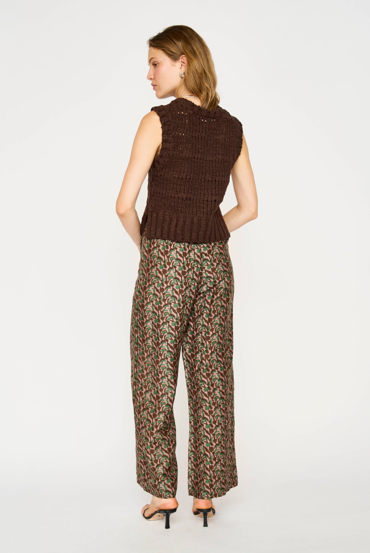 A woman is shown from the back wearing a sleeveless brown knit vest and wide-leg trousers with a dense red, green, and beige floral pattern, paired with black high-heeled sandals, against a plain white studio background.