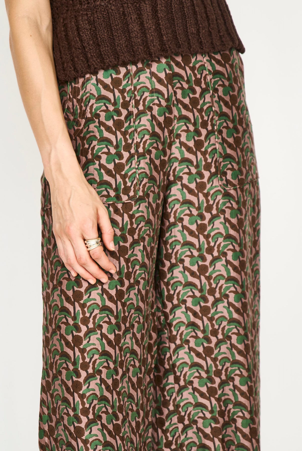 A close-up of a person wearing wide-leg trousers with an abstract brown and green pattern on a beige base, paired with a dark brown ribbed knit top against a plain light background.