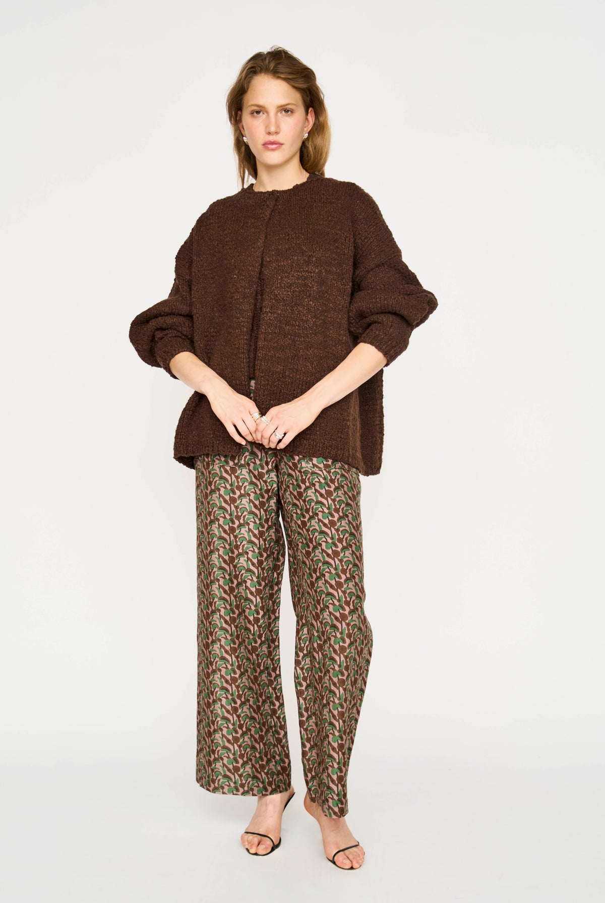 A woman modeling a brown textured knit cardigan and patterned wide-leg trousers with a red, green, and beige houndstooth-like print, standing against a light gray studio background.