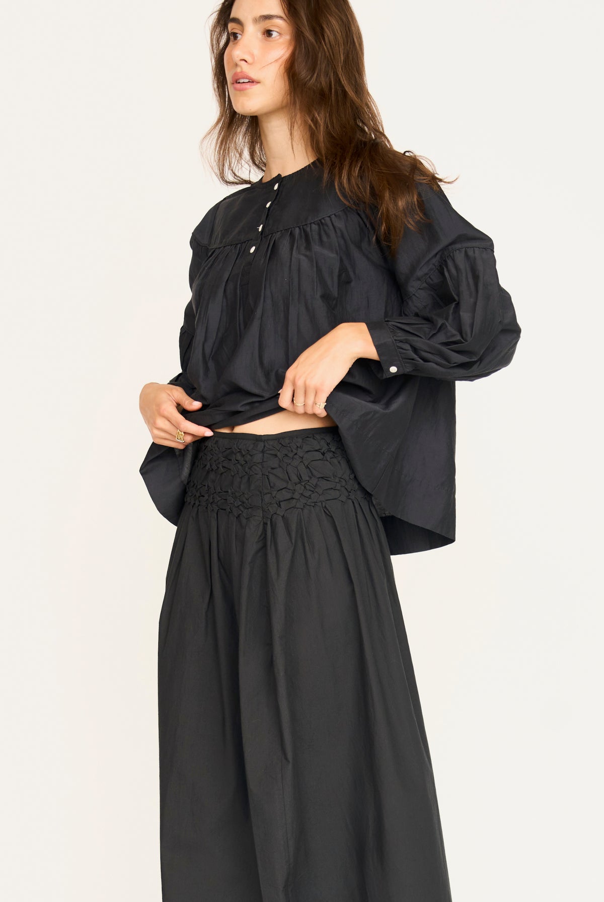 A woman modeling a black long-sleeve blouse with a gathered neckline and pleated sleeves, paired with black wide-leg pants, against a plain light background.