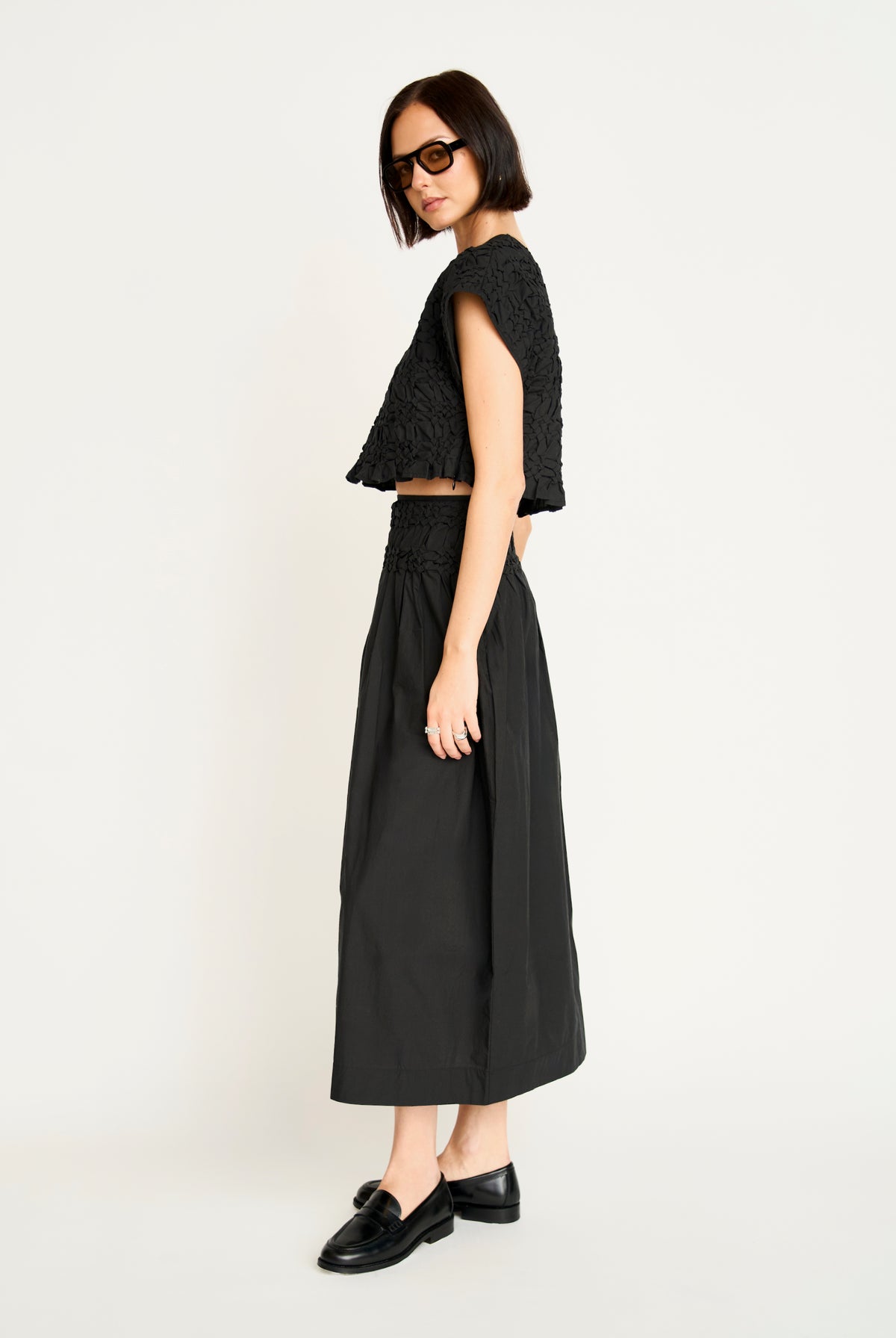 A model wearing a black textured sleeveless crop top and a matching black high-waisted midi skirt, styled with black loafers and sunglasses, against a plain off-white studio background.