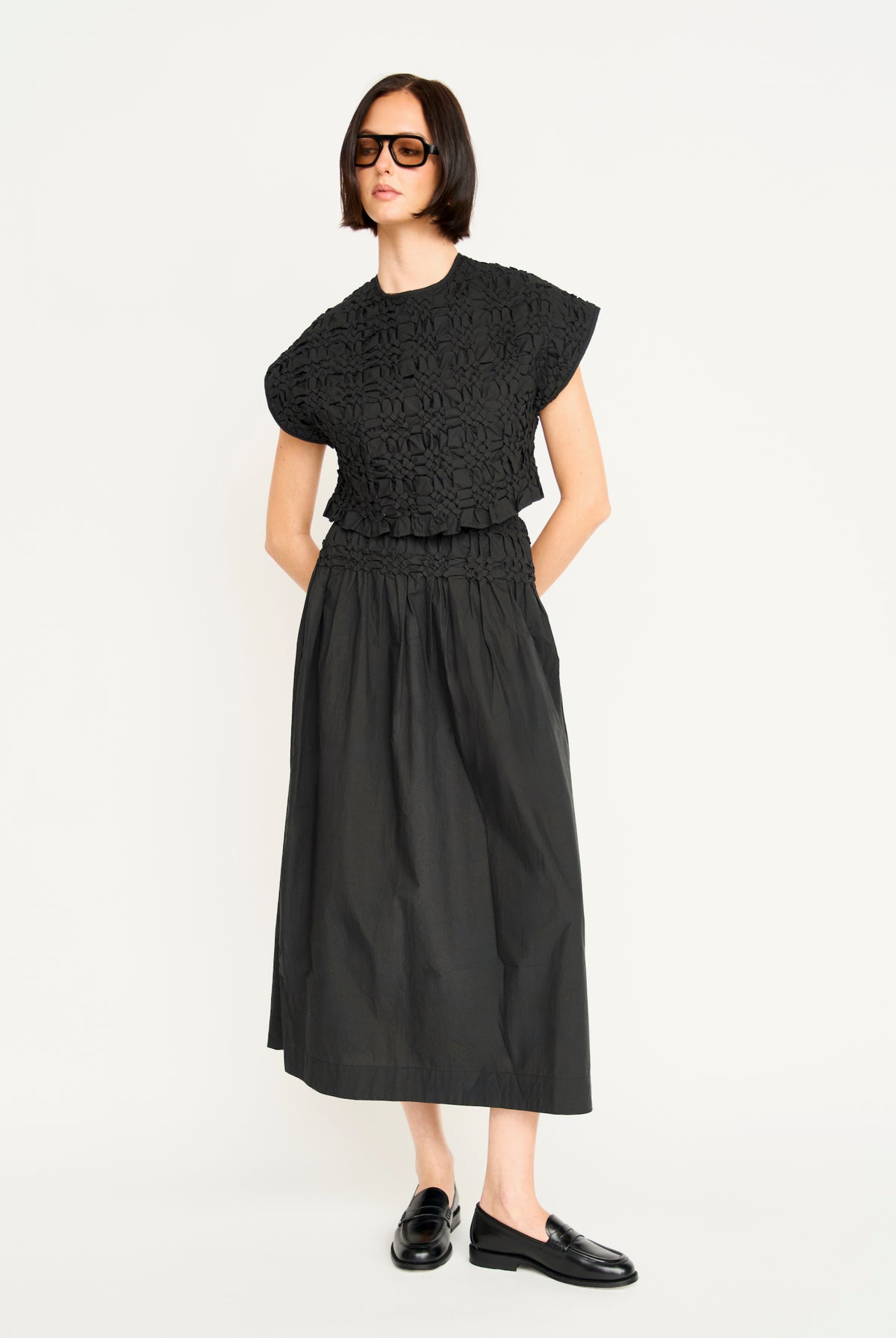 A model wearing a black two-piece outfit consisting of a short-sleeve cropped top with a textured floral lace overlay and a matching black midi skirt, paired with black leather loafers against a plain white studio background.