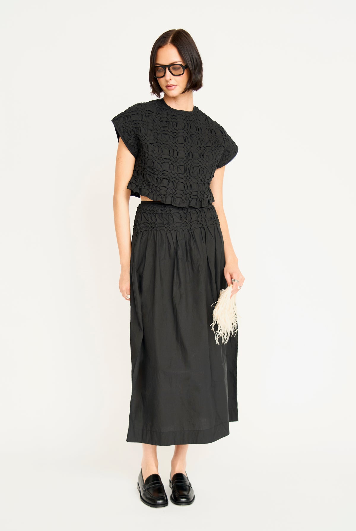 A model wearing a black two-piece outfit consisting of a short-sleeve cropped knit top and a matching midi skirt, paired with black loafers and holding a white fringed clutch against a plain white background.