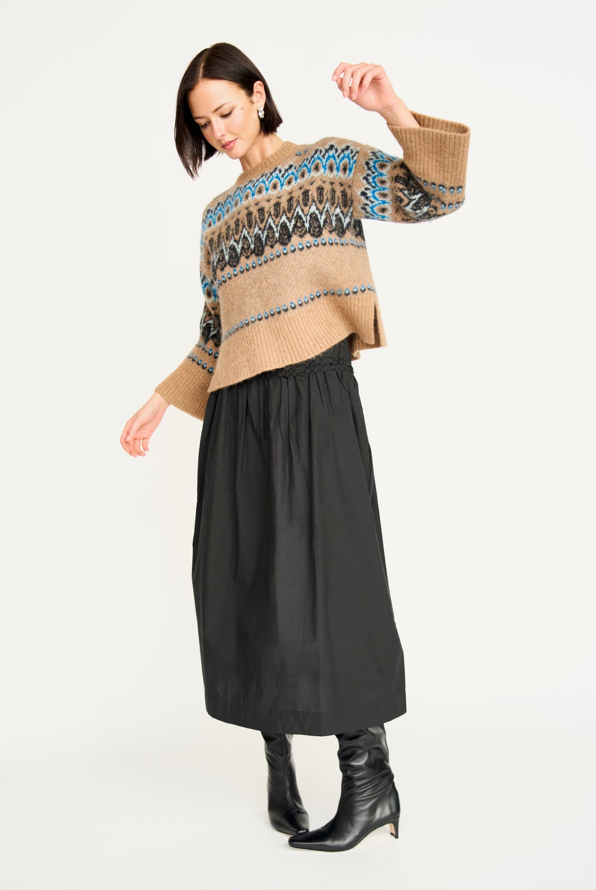 A woman modeling a cropped knit sweater with a beige base and a colorful Fair Isle pattern in blue, black, and brown, paired with a black midi skirt and black ankle boots, against a plain white studio background.