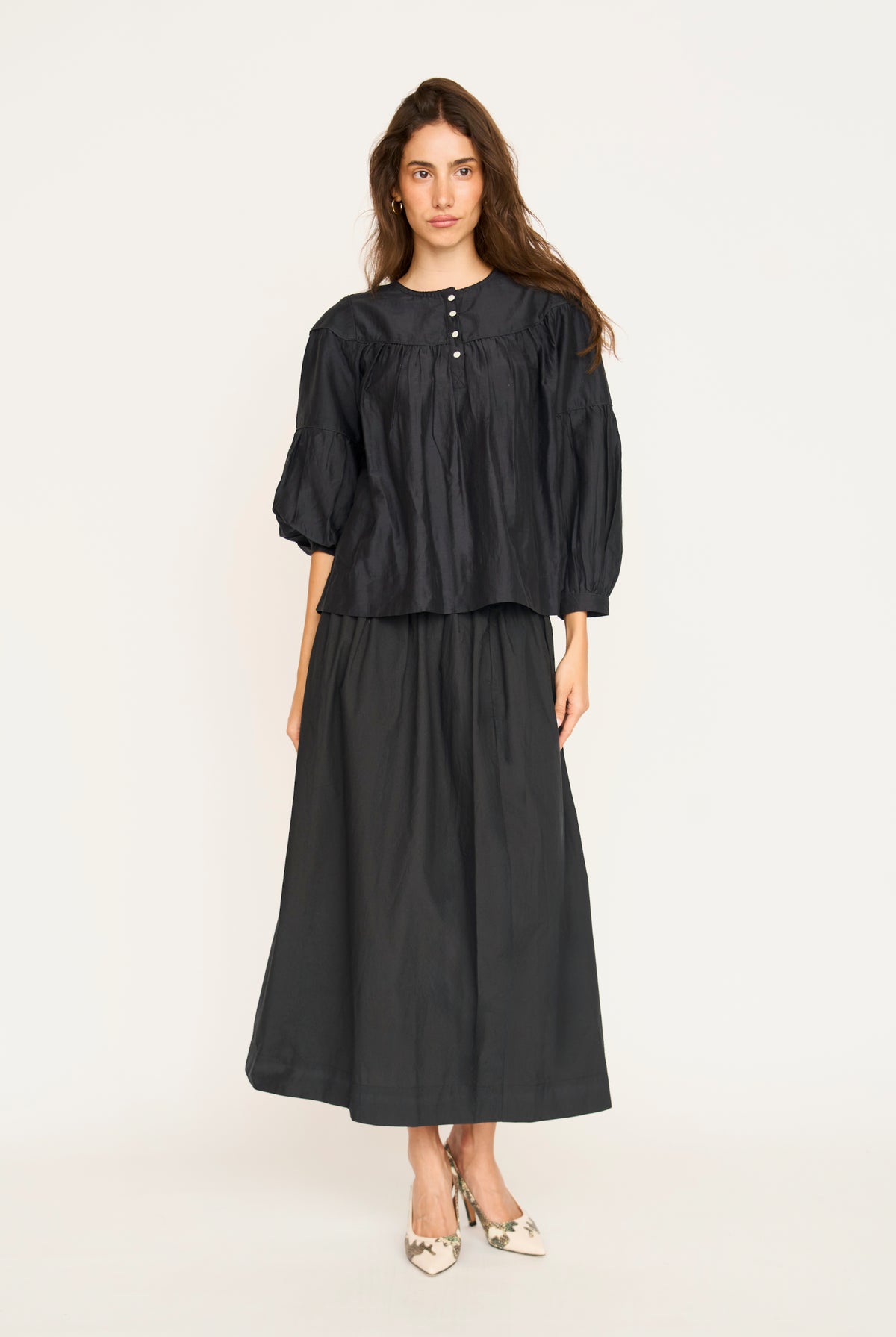A woman modeling a black two-piece outfit consisting of a short-sleeved, gathered blouse with a buttoned neckline and a matching high-waisted maxi skirt, paired with pointed shoes, against a plain white studio background.
