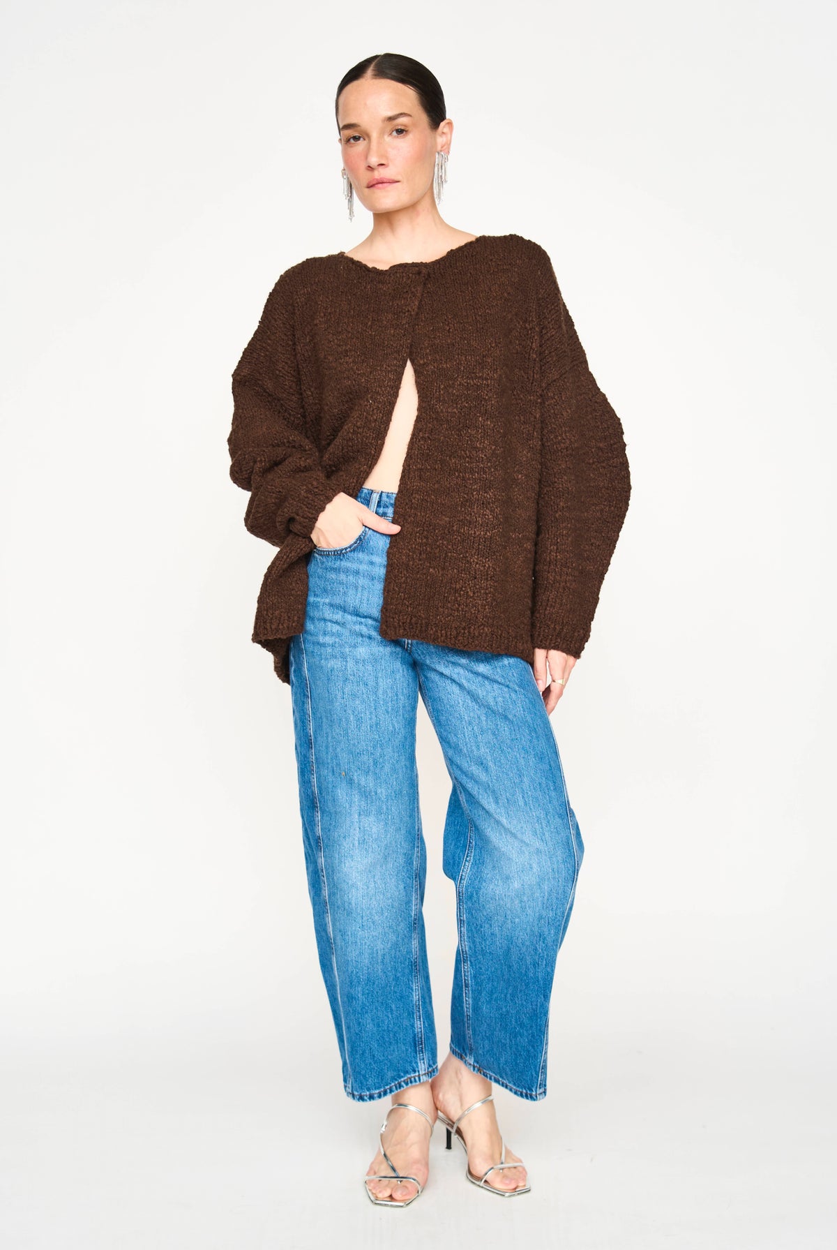 A woman modeling a dark brown textured knit sweater with a front slit, paired with wide-leg blue jeans and clear high-heeled sandals, against a plain white studio background.