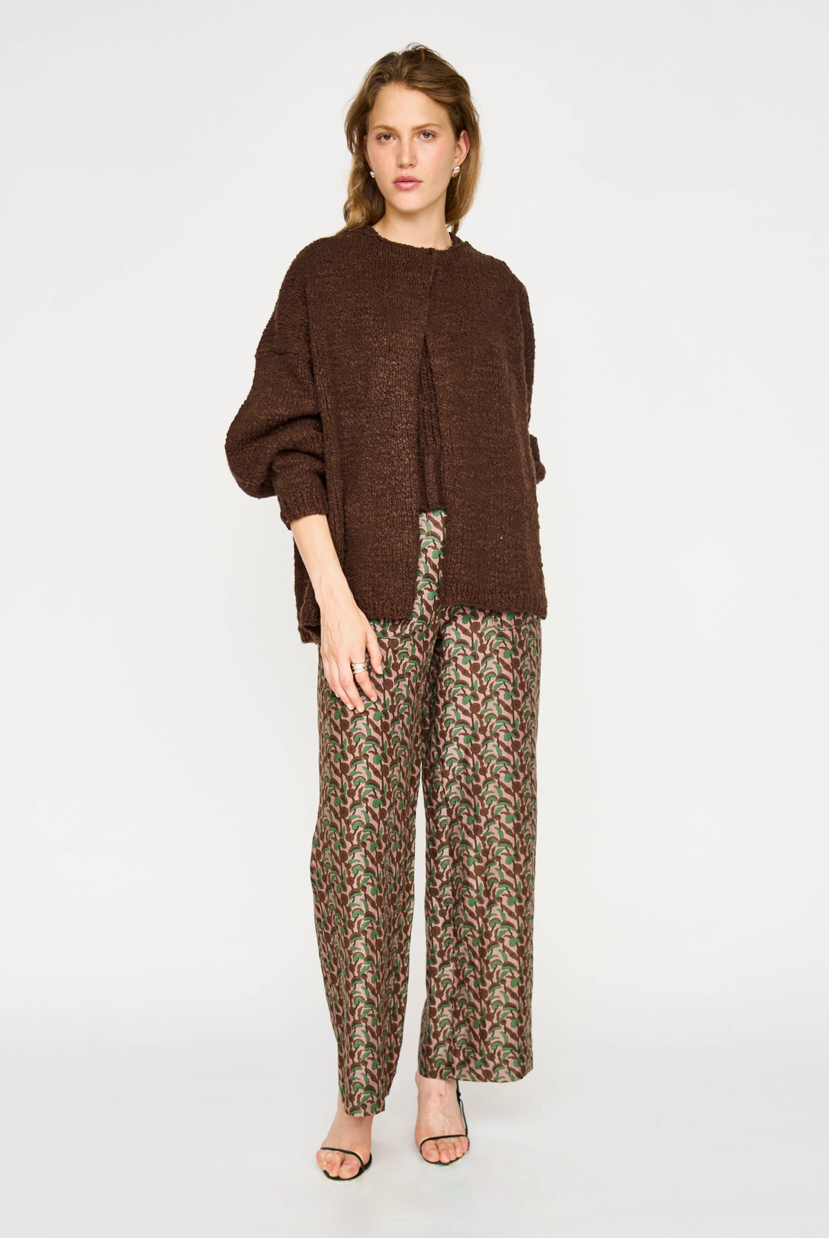 A model stands against a light gray studio background wearing a brown knit cropped cardigan, patterned wide-leg pants, and clear high-heeled sandals.
