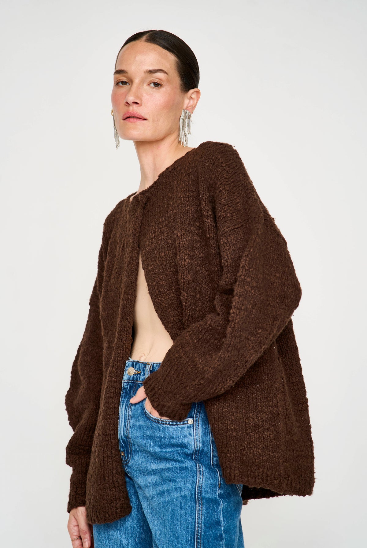 A woman wearing a dark brown fuzzy open-front cardigan over a beige top and blue jeans, photographed against a plain light gray studio background.