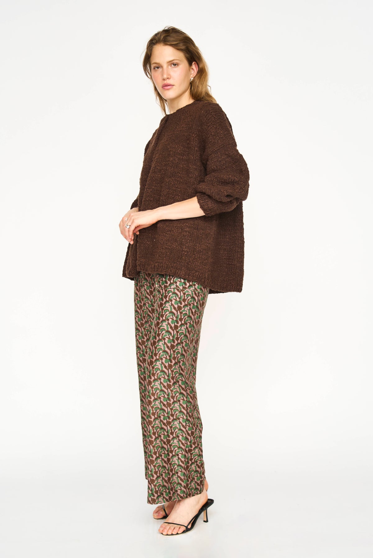 A woman modeling a brown textured knit cardigan and patterned wide-leg trousers with black heeled sandals, photographed against a plain white studio background.