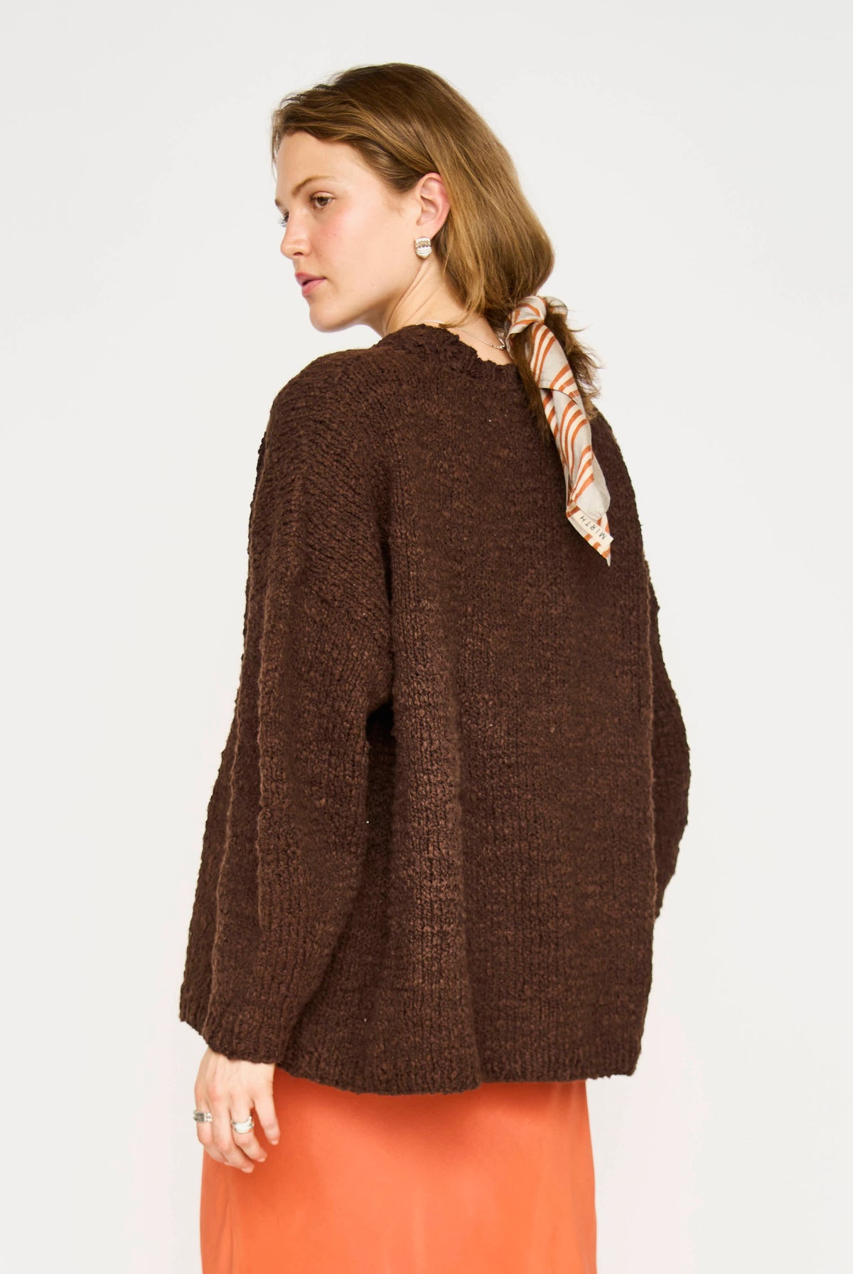 A woman shown from the back wearing a dark brown textured knit sweater and an orange skirt, with a patterned scarf tied around her neck, against a plain white background.