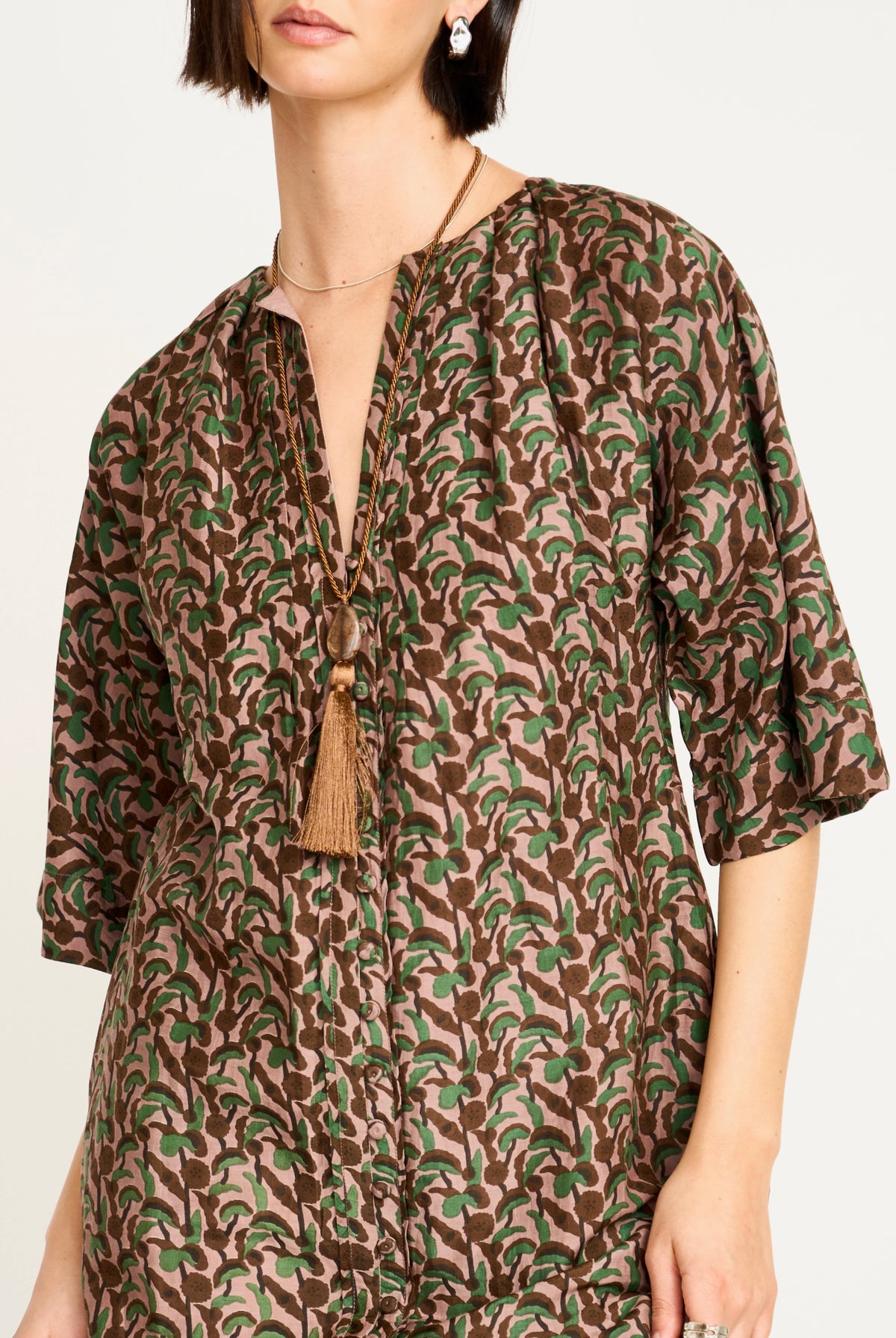 A woman wearing a brown and green abstract patterned tunic top with three-quarter sleeves and a tassel necklace, photographed against a plain light background.