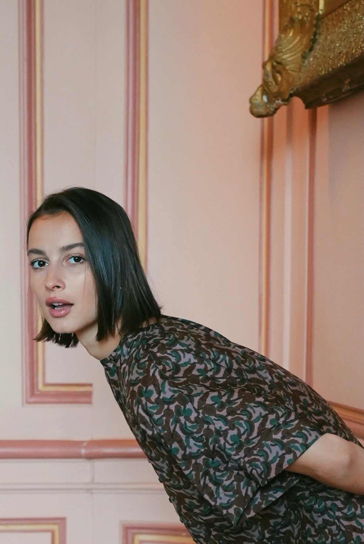 A woman with short dark hair leans in towards the camera, wearing a short-sleeve top with a dark brown and teal abstract pattern. She is posed in front of a pink paneled wall with gold trim.