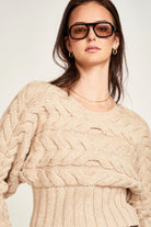 Woman wearing a beige cable knit sweater and sunglasses on a plain background