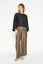 A woman modeling a black long-sleeve button-front blouse with wide cuffs, paired with brown and beige patterned wide-leg trousers, against a plain white studio background.