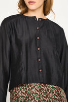 A close-up of a model wearing a black long-sleeve linen blouse with a buttoned neckline and a brown patterned skirt against a plain white background.