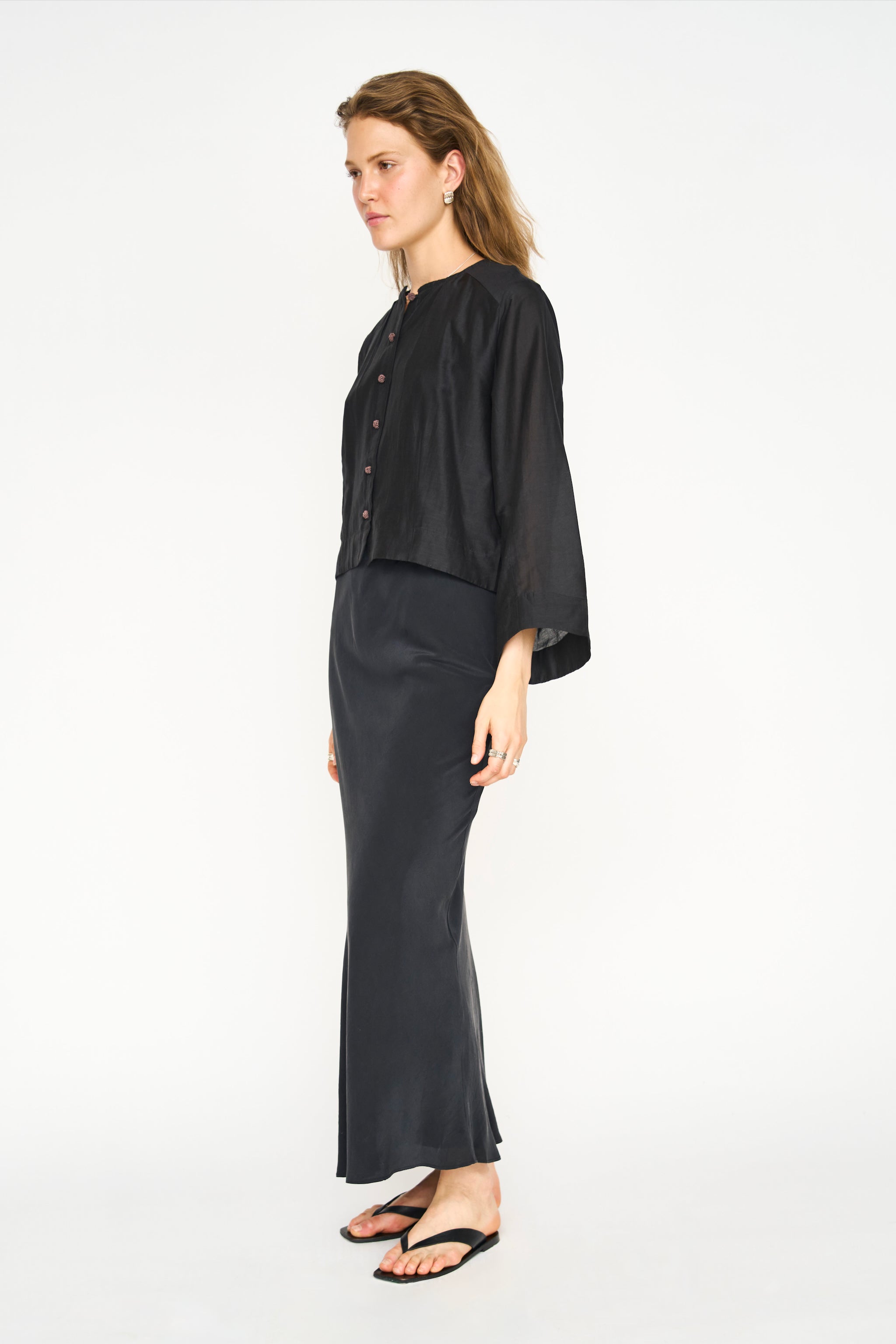 A woman modeling a black two-piece outfit consisting of a short-sleeved button-front top and a matching long skirt, photographed against a plain white studio background.